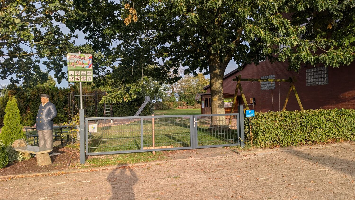 Eingang zum Mehrgenerationenplatz mit Spielplatzschild und der Seemannsfigur auf der Bank